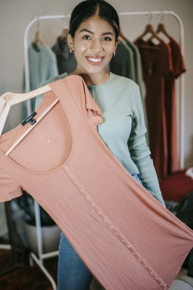 Confident ethnic woman in trendy outfit choosing clothes while demonstrating pink dress in light room at home and looking at camera happily